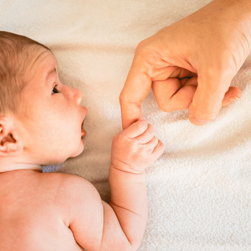 ein neugeborenes Baby liegt auf dem Bauch, schaut zur Seite und hält den Finger eines Erwachsenen