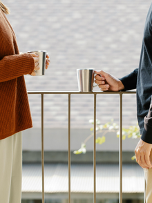 zwei menschen stehen sich an einem Geländer gegenüber. man sieht die Gesichter nicht, sie halten jeweils einen Becher in der Hand.